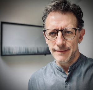 A headshot of Francisco J. Rivas Mesa, smiling in front of an artwork on a wall and wearing eyeglasses and a gray top.