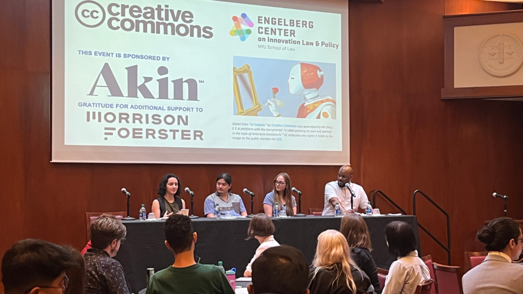 People seated at tables in a conference room watching a panel of four speak on stage below a slide with an image of a robot painting at an empty easel, saying: Creative Commons, Engleberg Center on Innovation Law & Policy, this event sponsored by Akin, gratitude for additional support to Morrison Foerster.